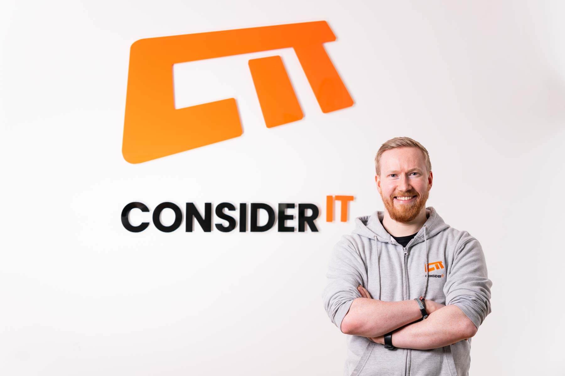 A picture of Rhys Smalridge one of Consider IT's IT Technicians standing by the Consider IT logo.