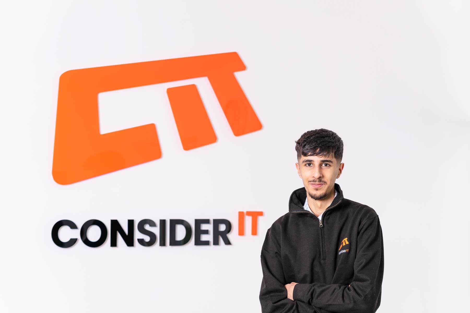 A picture of Rayaan Ali one of Consider IT's IT Technicians standing by the Consider IT logo.